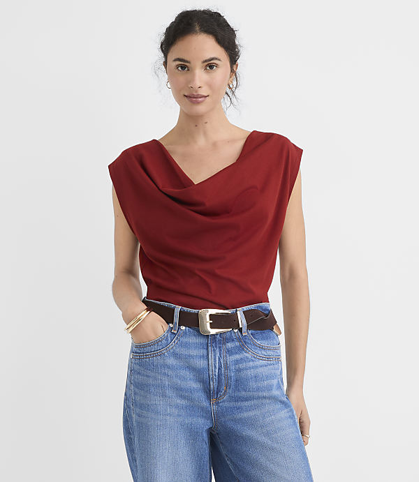 A woman in a rust-colored cowl-neck top and blue jeans stands against a plain white background.