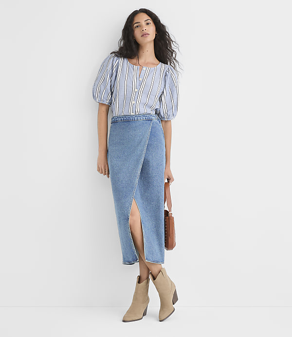 A woman in a striped blouse and denim skirt stands against a plain white background, holding a brown purse.