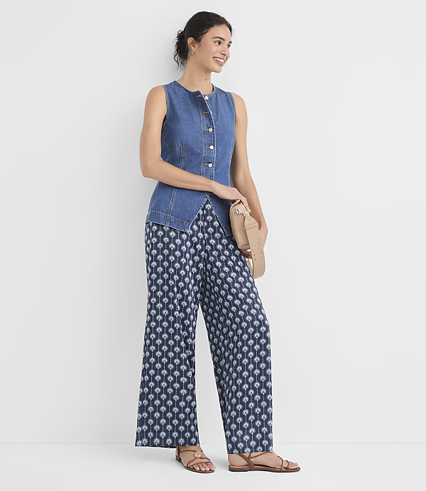 A woman in a denim vest and patterned pants stands against a plain white background, holding a tan clutch.