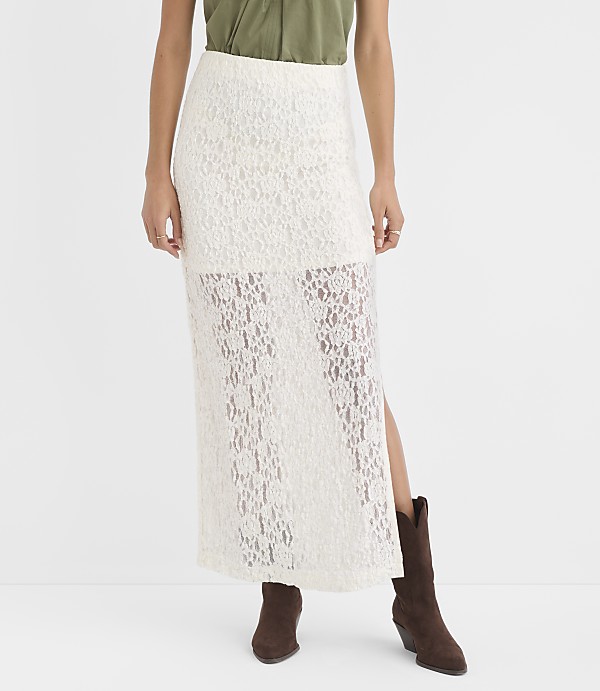 A woman in a white lace maxi skirt and brown boots stands against a plain white background.