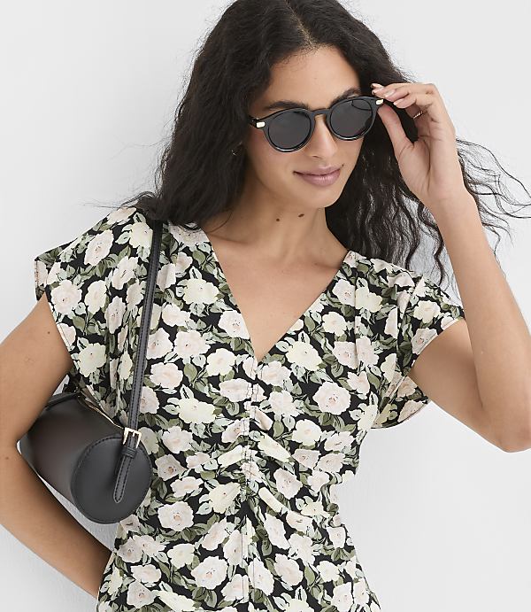 A woman in a floral dress and sunglasses adjusts her eyewear while a black cylindrical bag rests on her shoulder against a blurred background.