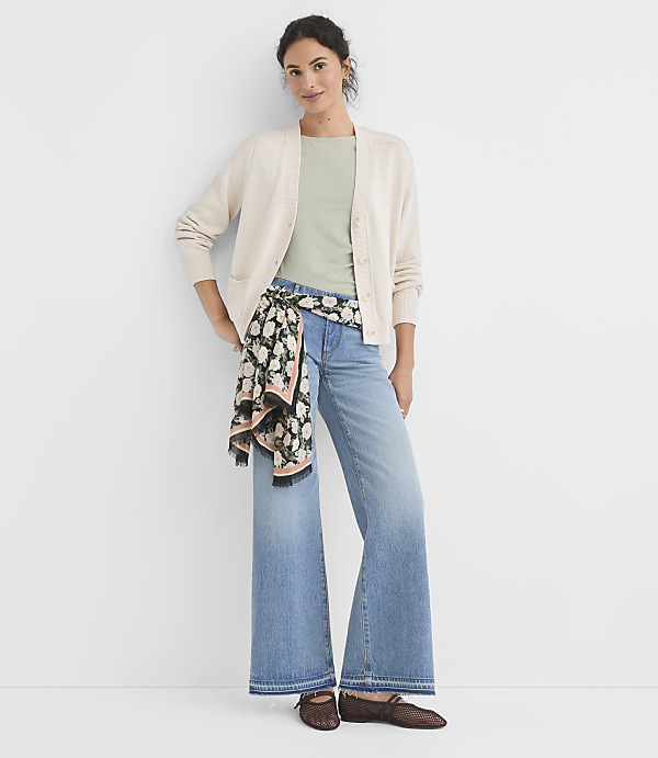 A woman in a cream cardigan and light blue wide-leg jeans stands against a plain white background, with a floral scarf tied around her waist.