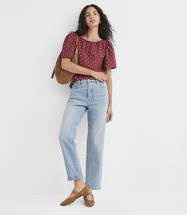 A woman in a patterned red top and light-wash jeans stands against a plain white background, carrying a brown bag.