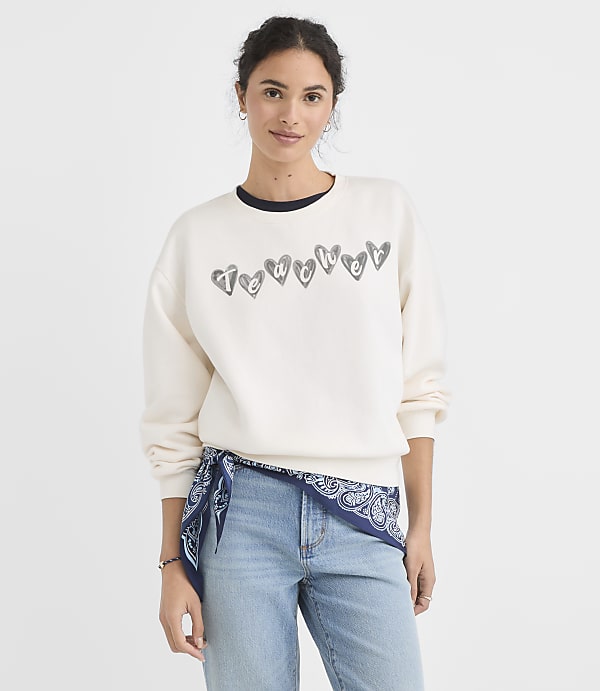 A woman in a cream sweatshirt with heart graphics and a blue bandana tied around her waist stands against a plain white background.
