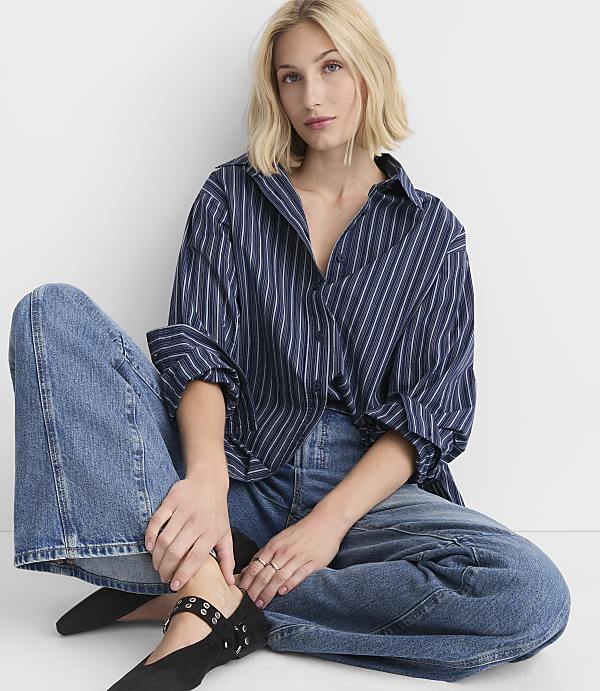 A blonde woman in a striped shirt and wide-leg jeans sits on a white floor against a white background.