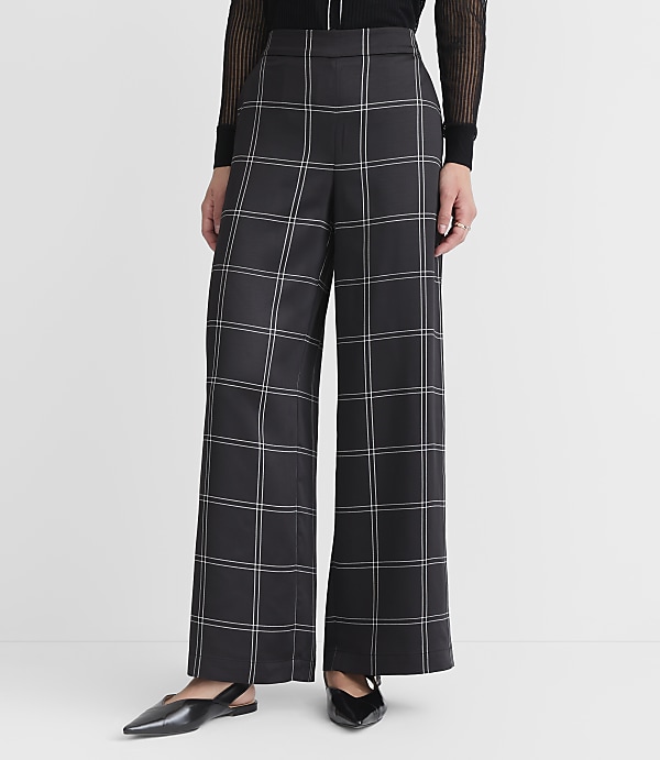 A person wearing black and white plaid wide-leg pants and black pointed-toe flats stands against a plain white background.
