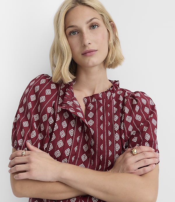A blonde woman with her arms crossed wears a patterned maroon dress against a plain white background.