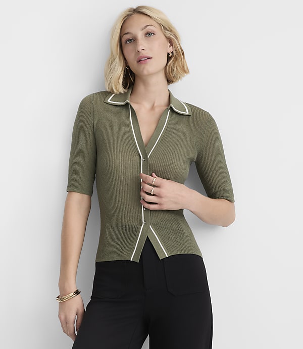 A woman in a green collared shirt and black pants stands against a plain white background.