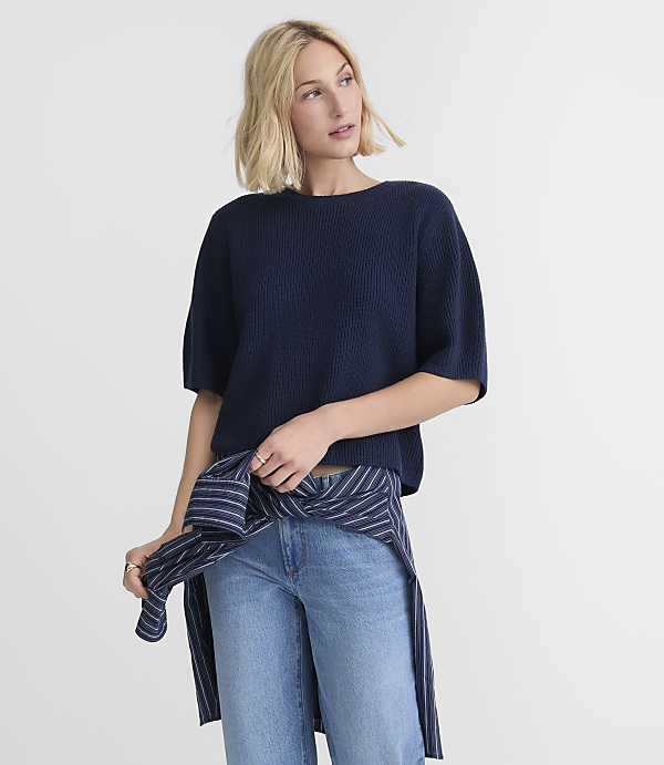 A blonde woman in a navy sweater and jeans stands against a plain white background, with a striped shirt tied around her waist.