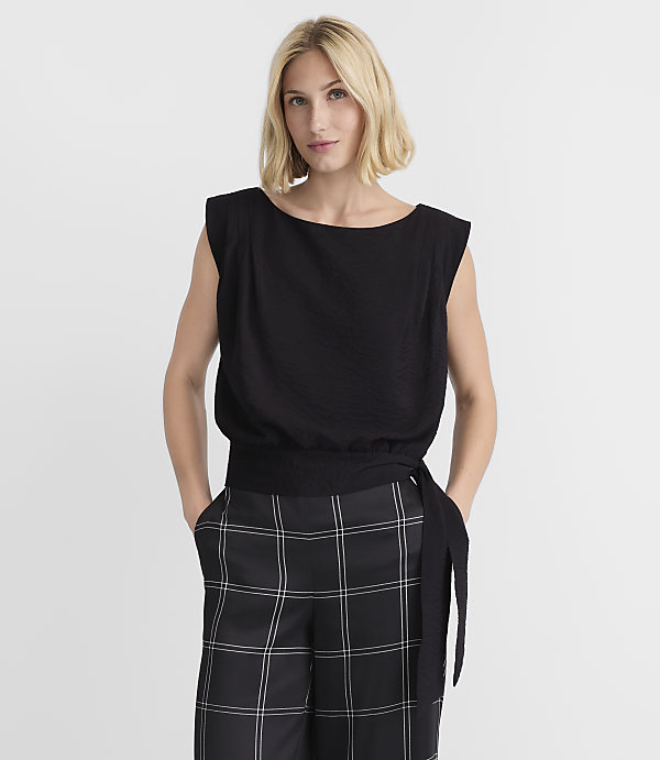 A blonde woman in a black top and plaid pants stands against a plain white background.