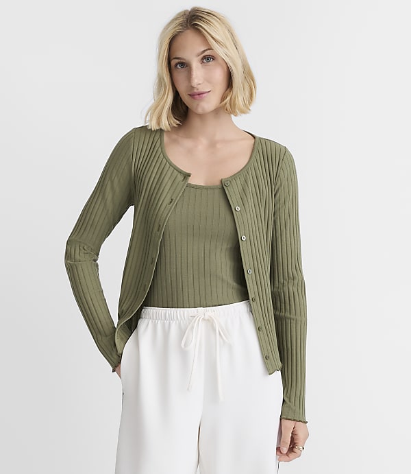 A blonde woman in a green ribbed cardigan and tank top stands against a plain white background.