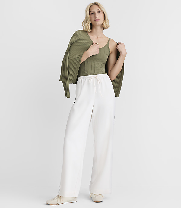 A woman in a green tank top and white pants stands against a plain white background, with a green cardigan draped over her shoulders.