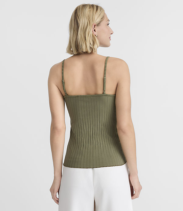 A woman in a green ribbed tank top and white pants stands against a plain white background.