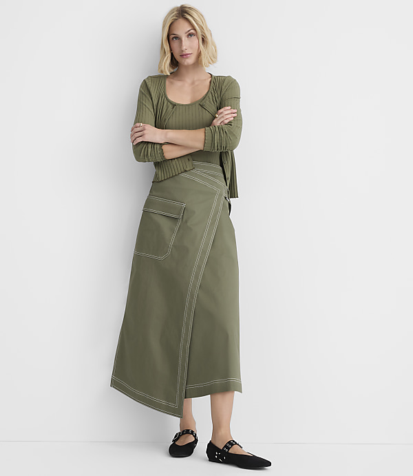 A woman in a green ribbed top and olive wrap skirt stands with her arms crossed against a plain white background.