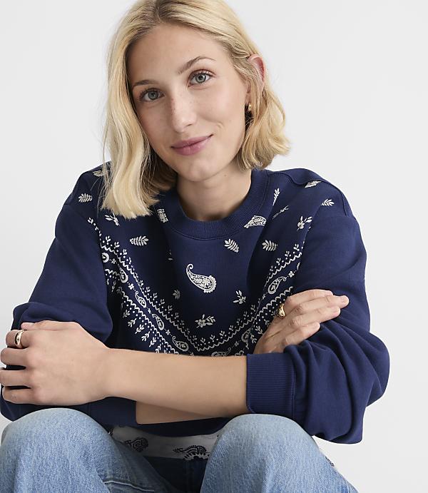 A blonde woman in a navy bandana-print sweatshirt and jeans smiles at the camera against a plain white background.