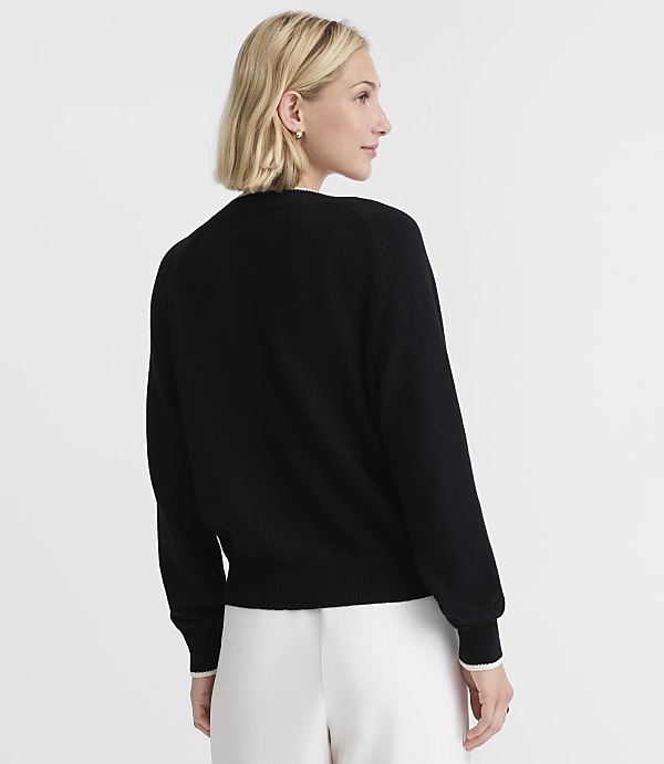 A woman in a black sweater and white pants stands against a plain white background.