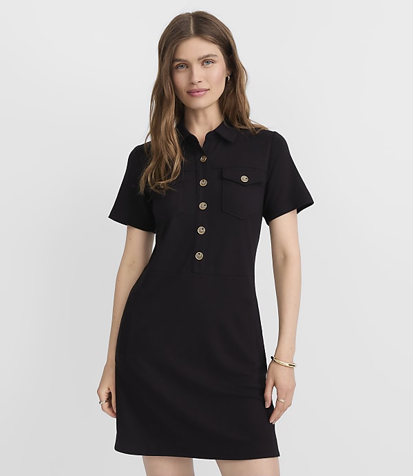 A woman in a black collared dress with gold buttons stands against a plain gray background.