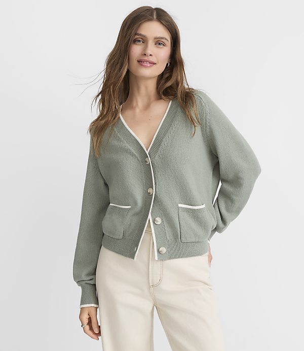 A woman in a sage green cardigan with white trim stands against a plain, light gray background.