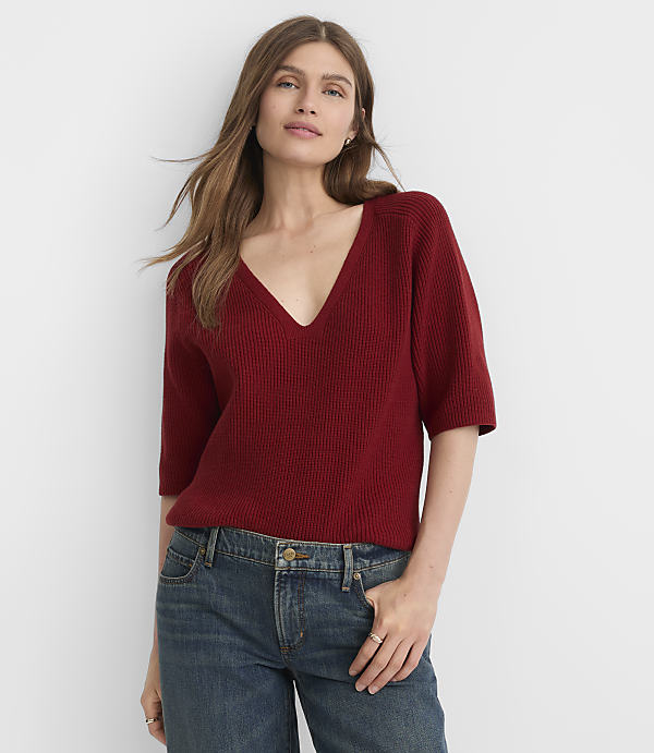 A woman in a red v-neck sweater and blue jeans stands against a plain white background.