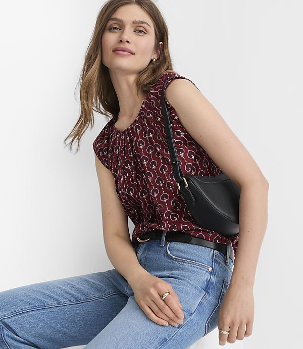 A woman in a patterned maroon top and blue jeans poses with a black purse against a plain white background.