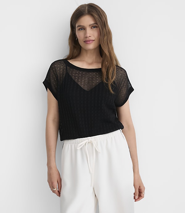 A woman in a black sheer top and white pants stands against a plain white background.