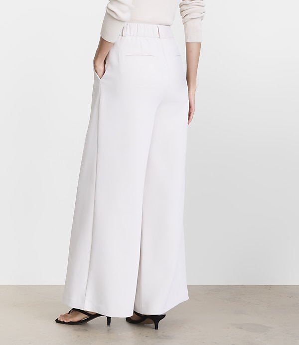 A person wearing white wide-leg pants and black heels stands against a plain, light-colored background.