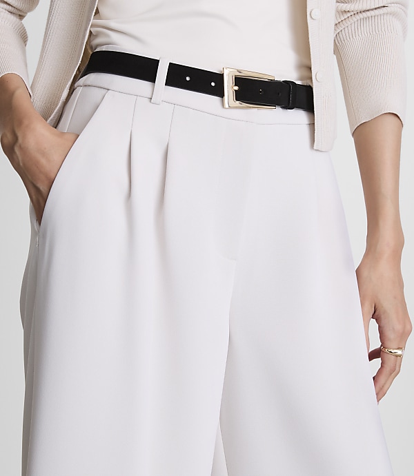 A person's hands are in the pockets of light-colored wide-leg pants, cinched with a black belt, against a blurred background of a cream cardigan.