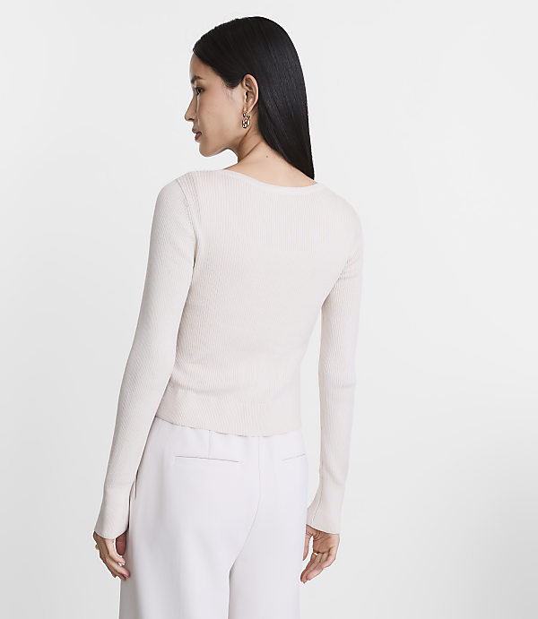 A woman in a cream-colored ribbed sweater and white pants stands against a plain white background.