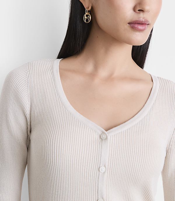 A woman with dark hair and gold earrings wears a cream-colored ribbed cardigan against a plain white background.