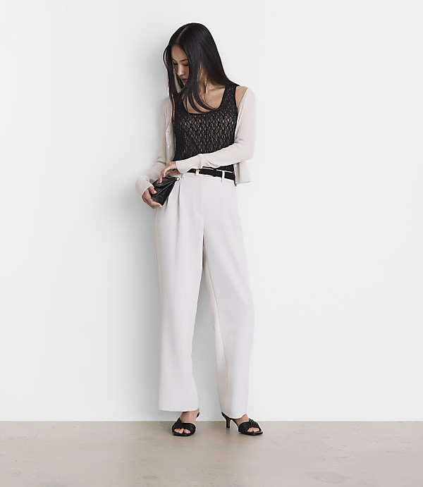 A woman in a black lace top, white pants, and a light cardigan stands against a plain white wall, holding a small black clutch.
