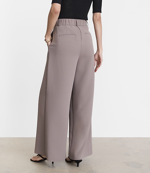 A person wearing wide-leg taupe pants and a black top stands with their back to the camera against a plain white background.