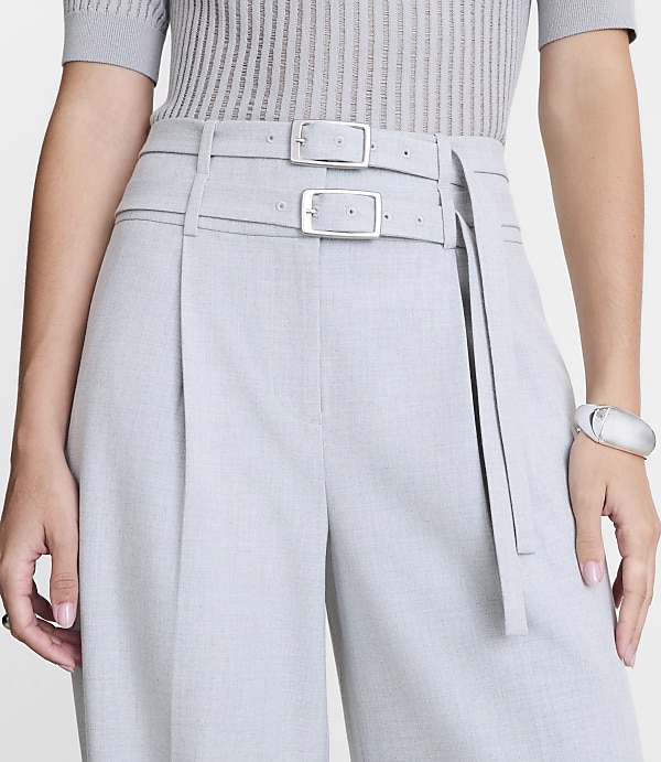 A person in a grey ribbed top and light grey pleated pants with a double buckle belt stands against a plain white background.