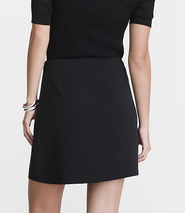 A person in a black top and skirt stands against a plain white background, with a silver bracelet on their left wrist.