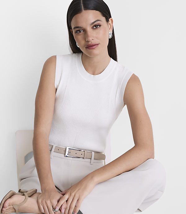 A woman in a white ribbed tank top and beige pants sits on a chair against a plain white background.