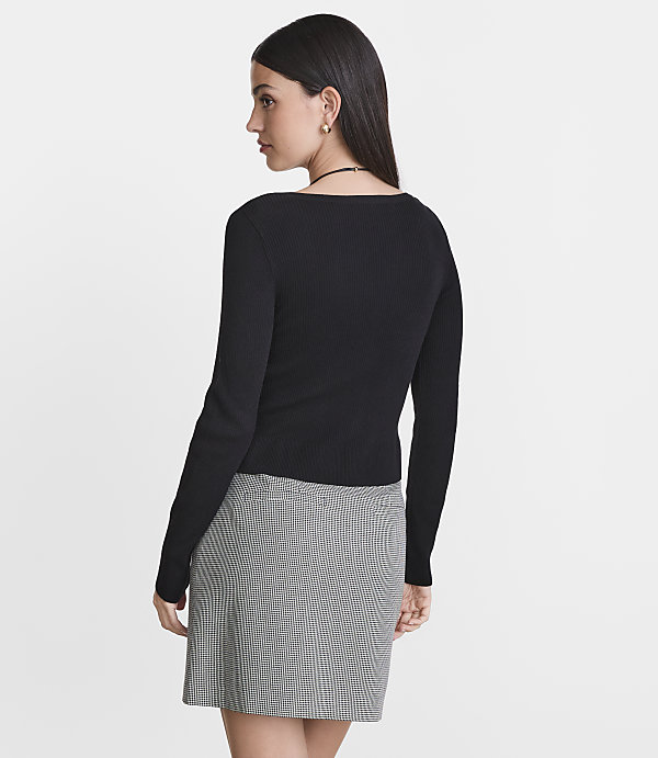 A woman in a black ribbed top and a checkered skirt stands against a plain white background.