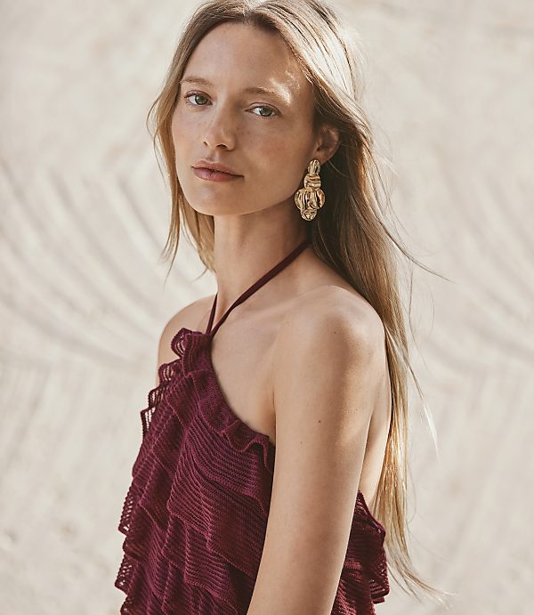 A woman with long blonde hair and large gold earrings wears a ruffled maroon halter top, with a sandy, palm-leaf-patterned background.