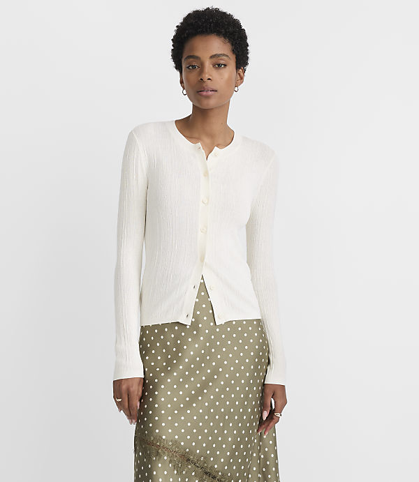 A woman in a cream-colored cardigan and a polka-dot skirt stands against a plain white background.