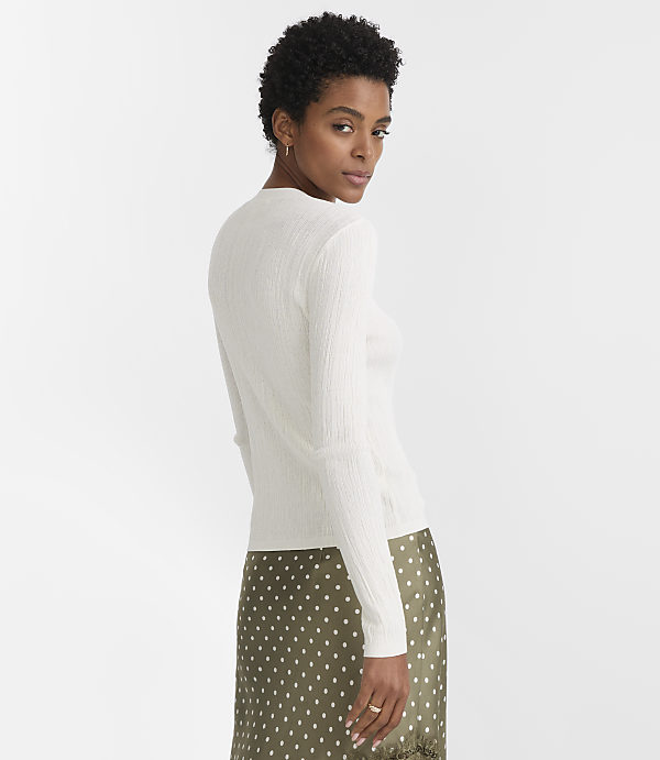 A woman in a white ribbed sweater and a green polka-dot skirt stands against a plain white background.