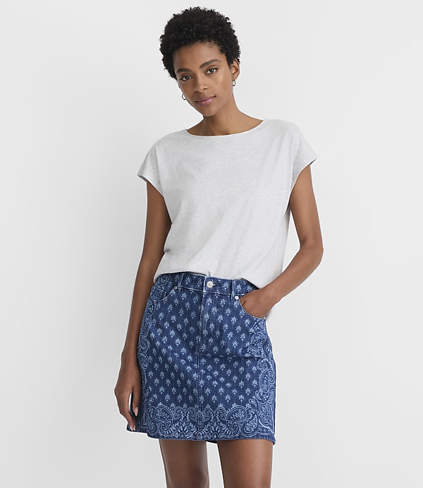A woman in a grey t-shirt and a patterned denim skirt stands against a plain white background.