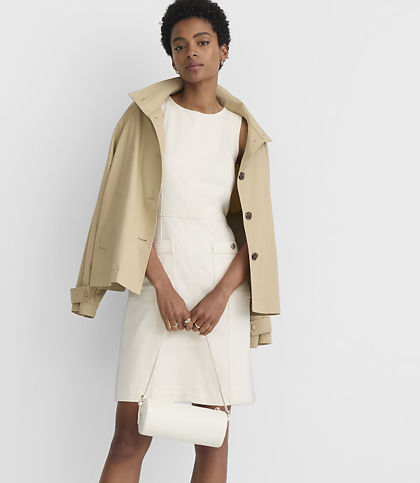 A woman in a cream dress and tan jacket holds a white cylindrical purse in front of a plain white background.