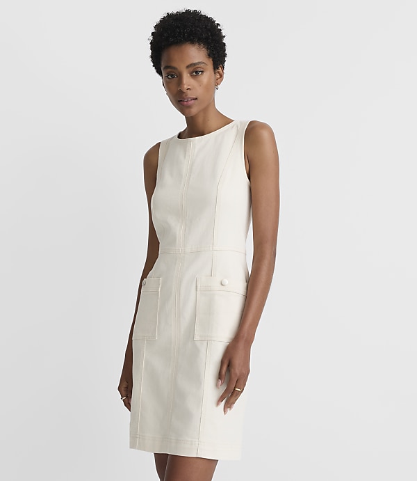 A woman in a cream-colored sleeveless dress with prominent stitching and pockets stands against a plain, light gray background.