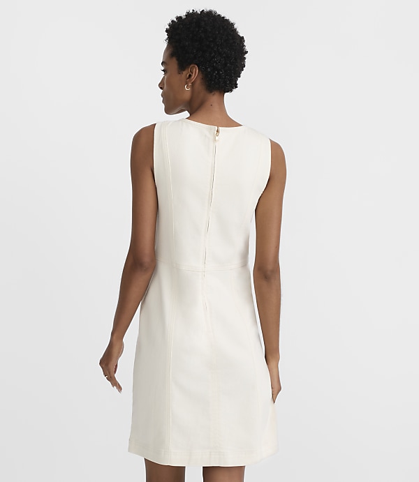 A woman in a cream-colored sleeveless dress stands against a plain white background.