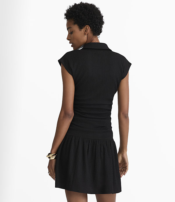 A woman in a black ruched dress stands against a plain white background.
