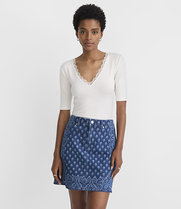 A woman in a white lace-trimmed top and a patterned denim skirt stands against a plain white background.