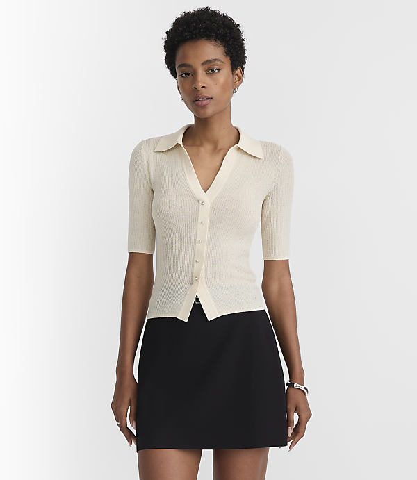 A woman in a cream-colored collared shirt and black skirt stands against a plain white background.