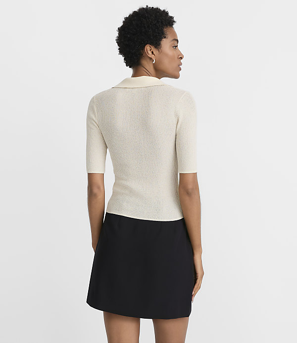 A woman in a cream-colored ribbed polo shirt and black skirt stands against a plain white background.