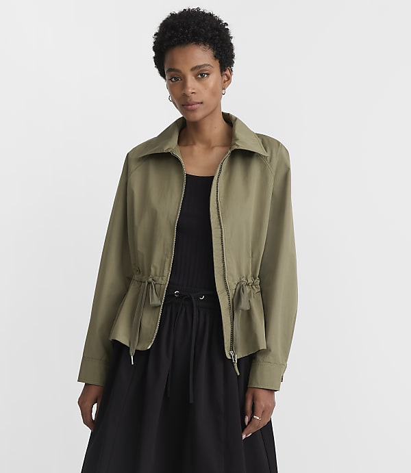 A woman in a green jacket and black skirt stands against a plain white background.