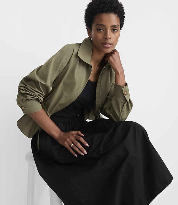 A woman in a green jacket and black skirt sits on a white stool against a plain white background.