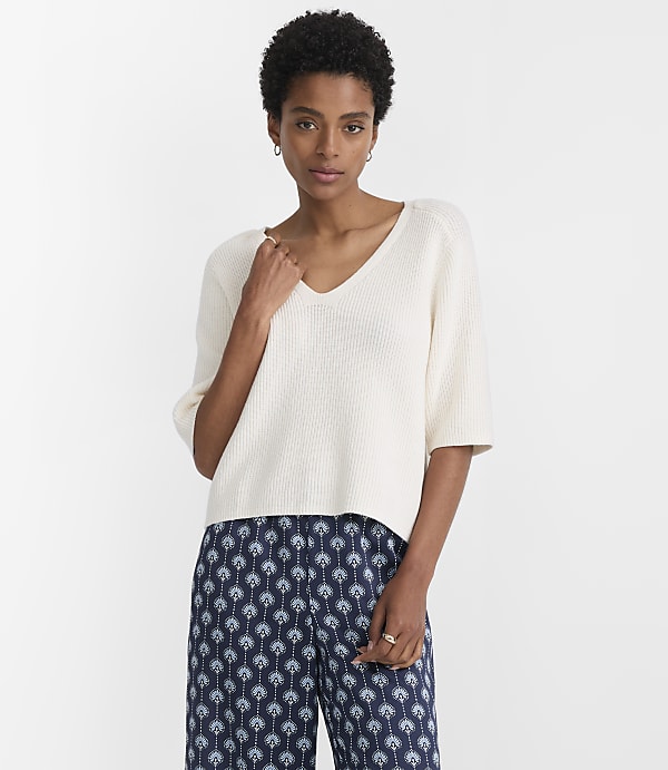 A woman in a cream-colored v-neck sweater and patterned blue pants stands against a plain white background.