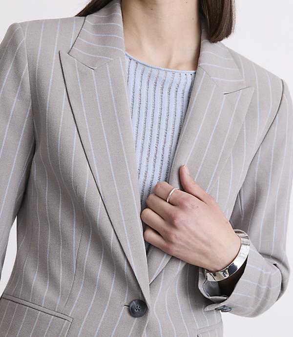 A woman in a pinstripe blazer and light blue top poses with her hand on her chest, against a plain background.